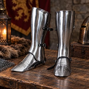 May include: A pair of polished steel greaves and sabatons designed to protect the lower legs and feet. These metallic armor pieces feature articulated foot plates, smooth curved shin guards, and black leather straps with metal buckles for fastening.