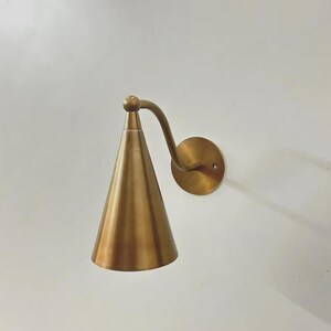 May include: A wall-mounted light fixture featuring a conical brass shade suspended from a curved arm attached to a circular brass wall plate. The metal has a smooth, polished finish with a warm golden tone, creating a sleek and functional design.