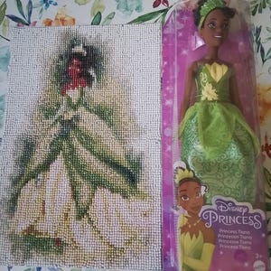 Può includere: Un progetto di pittura con diamanti che raffigura un personaggio in un abito verde accanto a una bambola Disney Princess Tiana in scatola. La bambola indossa un abito verde con corpetto giallo e fascia abbinata, confezionata in una scatola viola con il testo Disney Princess.