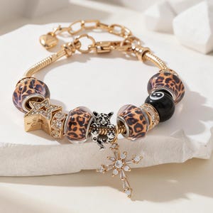 Op de afbeelding: Een goudkleurige bedelarmband met kralen in een bruin en zwart luipaardpatroon, een zwarte achtbal-kraal, een gouden sterbedel met heldere kristallen, een zwart-witte panterbedel en een met kristallen ingelegde kruisvormige hanger.