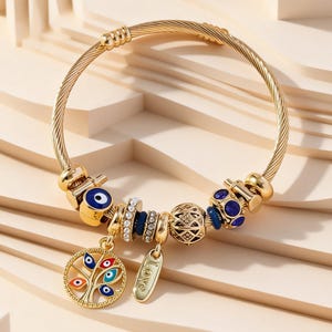 May include: A gold-tone cable wire bangle bracelet featuring a variety of charms, including a circular tree of life pendant with colourful evil eye motifs, a rectangular tag engraved with the word LOVE, and several beads with blue and clear accents.
