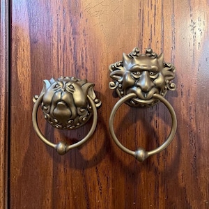 May include: Two antique-style brass door knockers mounted on a wood surface. The left knocker features a bulldog face with a circular ring, while the right knocker depicts a horned, mythical creature face with a larger circular ring attached.