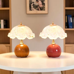 Può includere: Due lampade da tavolo con paralumi in vetro smerigliato smerlato con macchioline dorate. Una lampada ha una base rotonda con venature del legno, l'altra una base sferica rossa lucida. Entrambe le lampade presentano finiture in metallo dorato.