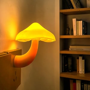 May include: A mushroom-shaped plug-in night light emitting a warm yellow glow. The lamp features a curved stem and a wide, rounded cap that illuminates the surrounding wall. It is directly connected to a wall outlet, providing soft ambient lighting.