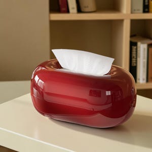 Op de afbeelding: Een glanzende, dieprode, ovaalvormige tissueboxhouder met een glad, reflecterend oppervlak. Een enkel wit tissue is door de centrale rechthoekige opening aan de bovenkant getrokken en staat rechtop tegen de afgeronde, pilvormige container aan.