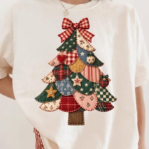 May include: An ivory short-sleeved t-shirt featuring a large graphic of a Christmas tree made of various patterned fabric patches in shades of red, green, blue, and yellow, topped with a red and white checkered bow and small ornaments.