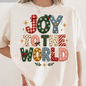 May include: An ivory t-shirt featuring a multi-colored graphic with the text JOY TO THE WORLD. The letters are designed with various patterns including red and white gingham, green floral, blue denim, and plaid, accented with small bows and stars.