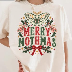May include: An ivory t-shirt featuring a graphic print with the text MERRY MOTHMAS in red and green. The design includes a large central moth, two smaller moths, holly leaves, berries, and a red bow, all arranged in a festive holiday pattern.