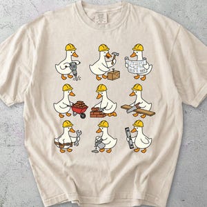 Op de afbeelding: Een ivoorkleurig t-shirt met een grafische print van negen witte eenden die gele bouwhelmen dragen. Elke eend is afgebeeld terwijl deze een andere taak uitvoert met gereedschap, waaronder een drilboor, hamer, bouwtekeningen, kruiwagen, bakstenen, zaag, moersleutel, emmer en waterpas.