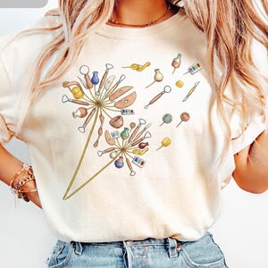 May include: An ivory t-shirt featuring a graphic print of two dandelion-like shapes constructed from various pottery and sculpting tools, including wooden ribs, wire loops, small ceramic bowls, and paint palettes, with scattered individual tools.