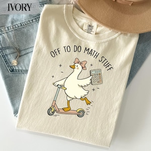 Könnte beinhalten: Ein elfenbeinfarbenes T-Shirt mit einer Grafik einer weißen Gans mit rosa Schleife, die auf einem Roller fährt und einen Taschenrechner hält. Der Text über der Gans lautet "OFF TO DO MATH STUFF" mit kleinen mathematischen Symbolen.