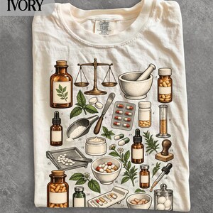 Könnte beinhalten: Ein elfenbeinfarbenes T-Shirt mit einem detaillierten Grafikdruck verschiedener Apothekerutensilien, darunter eine Waage, Mörser und Stößel, Glasflaschen, Pillenblister und Heilkräuter, die alle mit kleinen Nummern im wissenschaftlichen Stil versehen sind.