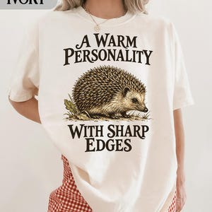 Op de afbeelding: Een ivoorkleurig t-shirt met een bruine en beige illustratie van een egel in het midden op de borst. De tekst boven de egel luidt A WARM PERSONALITY en de tekst eronder luidt WITH SHARP EDGES in een donkerbruin schreeflettertype.