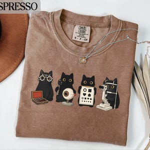 May include: A brown t-shirt featuring a graphic of four black cats performing eye exams with various optometry tools, including a trial frame, a model eyeball, an eye chart with shapes, and a slit lamp. The shirt has a Comfort Colors label.