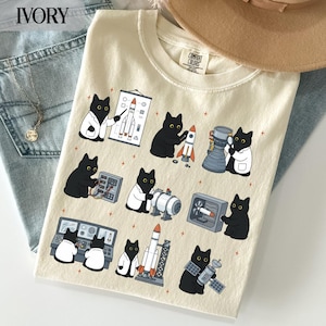 Puede incluir: Una camiseta de color marfil con un estampado gráfico de gatos negros vestidos como científicos e ingenieros trabajando con cohetes, equipos espaciales y paneles de control. La camiseta tiene cuello redondo y una etiqueta de la marca Comfort Colors.