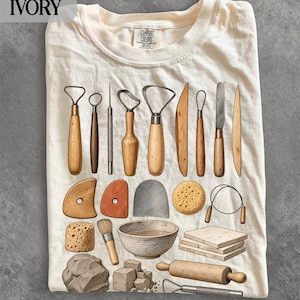 May include: An off-white t-shirt featuring a graphic print of various pottery tools, including wooden modelling sticks, wire loops, a sponge, a ceramic bowl, a stack of clay slabs, a rolling pin, and a lump of raw clay, with the text IVORY visible.