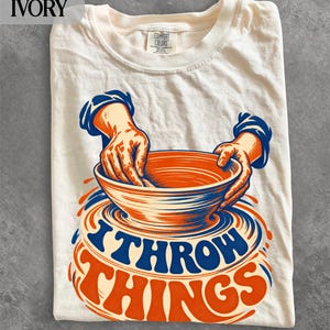 以下が含まれることがあります： アイボリー色のTシャツ。ろくろの上で粘土のボウルを成形する手のグラフィックが描かれています。オレンジと青の色使いで、ボウルの下に太字の曲線で「I THROW THINGS」というテキストが配置されています。Comfort Colorsのラベルが付いています。