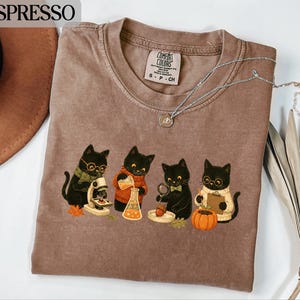 Könnte beinhalten: Ein braunes kurzärmeliges T-Shirt mit einer Grafik von vier schwarzen Katzen, die wissenschaftliche Experimente durchführen. Die Katzen tragen Pullover und Brillen und benutzen ein Mikroskop, Bechergläser und eine Lupe. Der Text ESPRESSO ist zu sehen.
