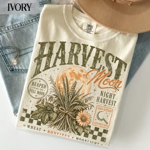 Könnte beinhalten: Ein elfenbeinfarbenes T-Shirt mit einem Grafikdruck mit dem Text Harvest Moon, Reaped Under The Full Moon, Night Harvest, Wheat, Bonfires und Moonlight. Das Design zeigt einen großen Mond, Weizenähren, eine Blume und ein Sichelsymbol.