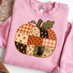Op de afbeelding: Een lichtroze sweater met ronde hals en een grote, centrale pompoenafbeelding die is samengesteld uit verschillende patchwork-stijl patronen, waaronder oranje gingham, bloemenprints, zwarte sterren, snoepmaïs, een zwarte kat en een spinnenweb.