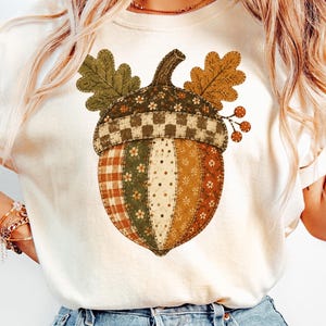 Op de afbeelding: Een crèmekleurig T-shirt met een grote afbeelding van een eikel. De eikel is samengesteld uit verschillende patchworkpatronen, waaronder ruiten, stippen en bloemenprints in bruin-, groen- en oranjetinten, met twee eikenbladeren aan de bovenkant.