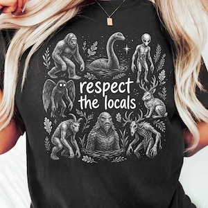 Może przedstawiać: Czarny t-shirt z białą grafiką przedstawiającą różne kryptydy, w tym Wielką Stopę, potwora z Loch Ness, kosmitę i jackalope'a. Napis "respect the locals" znajduje się na środku, otoczony szczegółowymi ilustracjami mitycznych stworzeń.
