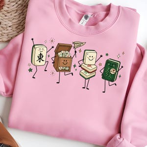 Pink sweatshirt featuring a cartoon of dancing mahjong tiles and a mahjong box.