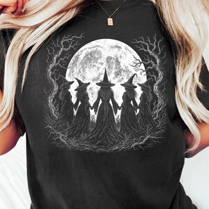 Könnte beinhalten: Ein schwarzes T-Shirt mit einem weißen Grafikdruck von fünf Gestalten in spitzen Hüten und langen Gewändern, die sich im Kreis an den Händen halten. Die Szene ist vor einem großen, texturierten Vollmond und kahlen, knorrigen Baumzweigen dargestellt.