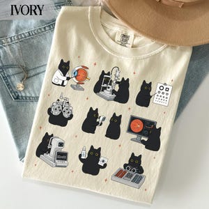 以下が含まれることがあります： アイボリー色のTシャツで、様々な眼科検査を行う黒猫のグラフィックプリントが施されています。イラストには、細隙灯顕微鏡、フォロプター、視力表などの医療機器を使用する猫たちが描かれており、小さなオレンジ色の星のアクセントが全体に散りばめられています。