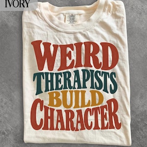 May include: An ivory t-shirt featuring the text WEIRD THERAPISTS BUILD CHARACTER in a bold, retro-style font. The words WEIRD and CHARACTER are printed in a deep red, THERAPISTS in dark teal, and BUILD in a mustard yellow colour.