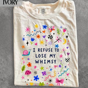 以下が含まれることがあります： アイボリー色のTシャツ。前面には「I REFUSE TO LOSE MY WHIMSY」という濃い青色の文字がプリントされ、その周囲にはカタツムリ、蝶、キノコ、花、星、ハート、幾何学模様などのカラフルな手描き風のイラストが描かれています。