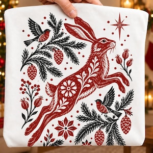 May include: A white fabric item featuring a red and black graphic print of a leaping hare surrounded by pine branches, pinecones, berries, and two small birds. The design includes floral patterns on the hare and a four-pointed star above it.