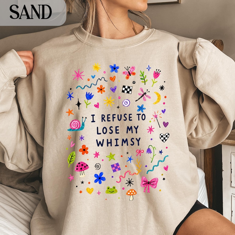 Sand colored sweatshirt with colorful cartoon illustrations and text "I REFUSE TO LOSE MY WHIMSY".