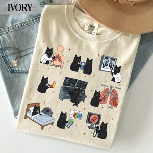 May include: A cream-coloured t-shirt featuring a graphic print of black cats performing medical procedures on lungs. The illustrations include cats using stethoscopes, reading X-rays, operating monitors, and using inhalers, with labels like Trachea.