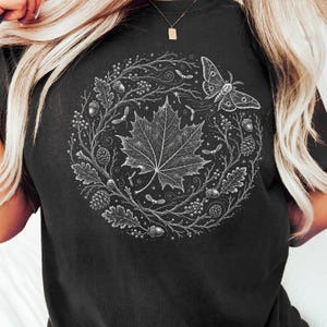 Op de afbeelding: Een zwart t-shirt met een witte cirkelvormige grafische print. Het ontwerp toont een centraal esdoornblad omringd door een krans van eikenbladeren, eikels, dennenappels, bessen en een mot, alles weergegeven in een gedetailleerde lijnstijl.