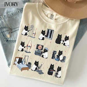 May include: An ivory t-shirt featuring a graphic print of black cats dressed as medical professionals performing various orthopaedic procedures, including examining x-rays, using medical equipment, and assisting patients in a clinical setting.