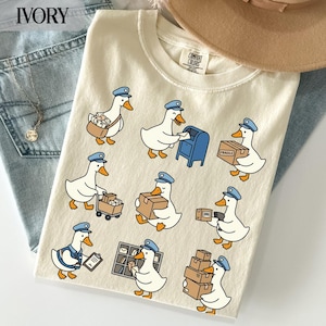 Könnte beinhalten: Ein cremefarbenes T-Shirt mit einem wiederkehrenden Muster aus weißen Cartoon-Enten, die als Postboten gekleidet sind. Die Enten tragen blaue Mützen und transportieren verschiedene Pakete, Umschläge und Postgegenstände wie einen blauen Briefkasten.