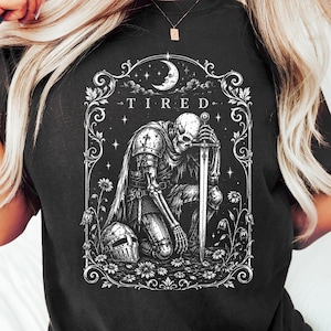Op de afbeelding: Een zwart t-shirt met een witte grafische print van een skelet-ridder in harnas die knielt en een zwaard vasthoudt. Het ontwerp bevat de tekst "TIRED" boven de ridder, omringd door een decoratieve bloemenrand, sterren en een maansikkel.