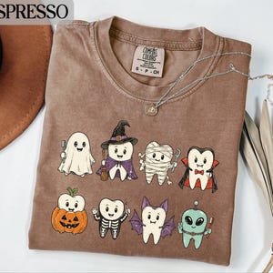Könnte beinhalten: Ein braunes Kurzarm-T-Shirt mit einem Grafikdruck von acht Cartoon-Zähnen in verschiedenen Halloween-Kostümen, darunter ein Geist, eine Hexe, eine Mumie, ein Vampir, ein Kürbis, ein Skelett, eine Fledermaus und ein Alien. Rundhalsausschnitt.
