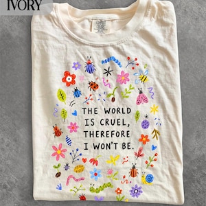 Op de afbeelding: Een ivoorkleurig t-shirt met een kleurrijke grafische print van diverse insecten, bloemen en bladeren rondom de zwarte tekst: THE WORLD IS CRUEL, THEREFORE I WON'T BE. Het t-shirt heeft een ronde hals en een Comfort Colors label in de kraag.