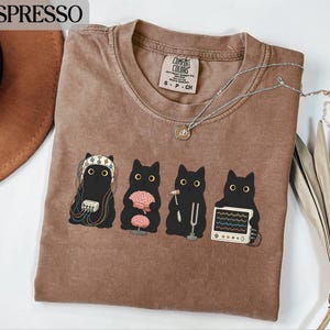 Puede incluir: Una camiseta de cuello redondo marrón con un gráfico de cuatro gatos negros realizando exámenes médicos. El primer gato lleva un gorro de EEG, el segundo sostiene un cerebro, el tercero un martillo de reflejos y un diapasón, y el cuarto un monitor.
