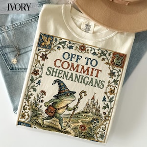 Op de afbeelding: Een ivoorkleurig t-shirt met een grafische print van een kikker met een tovenaarshoed met sterrenpatroon en een houten staf. De tekst luidt OFF TO COMMIT SHENANIGANS, omringd door een decoratieve bloemenrand met een kasteel en mythische wezens.