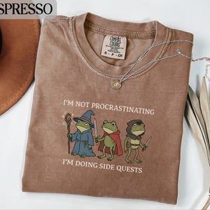 Könnte beinhalten: Ein braunes T-Shirt mit einer Grafik von drei als Fantasy-Figuren verkleideten Fröschen und dem Text I'M NOT PROCRASTINATING I'M DOING SIDE QUESTS. Das Shirt hat einen Rundhalsausschnitt, ein Etikett mit Größe S und eine silberne Halskette.