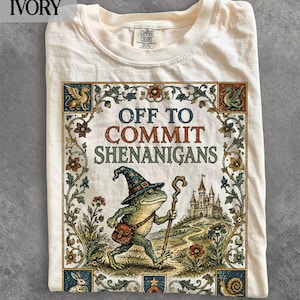 Puede incluir: Una camiseta color marfil con un estampado gráfico de una rana con un sombrero de mago con estrellas y un bastón de madera. El texto dice OFF TO COMMIT SHENANIGANS, rodeado por un borde floral decorativo, un castillo y animales pequeños.
