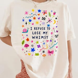 Ivory t-shirt with colorful whimsical drawings and text "I REFUSE TO LOSE MY WHIMSY".