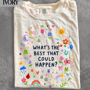 Könnte beinhalten: Ein elfenbeinfarbenes T-Shirt mit dem dunkelblauen Text "WHAT'S THE BEST THAT COULD HAPPEN?", umgeben von bunten, handgezeichneten Illustrationen wie Sonne, Mond, Regenbogen, Blumen, Sternen, Herzen, einem Marienkäfer, einem Schmetterling und Würfeln.