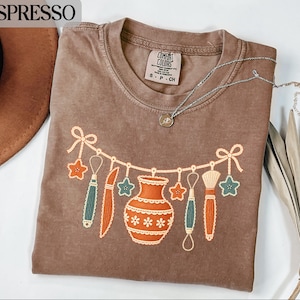 Peut inclure: Un t-shirt marron à manches courtes avec une impression graphique représentant une guirlande avec un vase orange central, divers outils de poterie et des formes d'étoiles. Le t-shirt a un col rond et une étiquette Comfort Colors.
