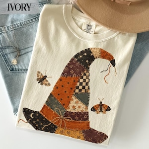 Op de afbeelding: Een ivoorkleurig t-shirt met een afbeelding van een patchwork heksenhoed in oranje, bruine en zwarte tinten met ster-, bloemen- en ruitpatronen. Het ontwerp bevat twee motten en een klein strikje van touw op de hoedband. Het shirt heeft een Comfort Colors label.