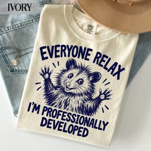 May include: An ivory-colored t-shirt featuring a dark blue graphic of an opossum with its paws raised. The text above the opossum reads "EVERYONE RELAX" and the text below reads "I'M PROFESSIONALLY DEVELOPED". The shirt has a crew neckline.