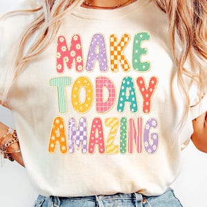 May include: A cream-coloured t-shirt featuring the text MAKE TODAY AMAZING in large, multi-coloured letters. Each letter has a unique pattern, including daisies, checkers, stripes, polka dots, and hearts in shades of pink, yellow, orange, green, and purple.